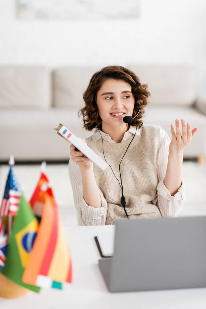 happy language teacher in headset holding French dictionary while talking during online lesson near international flags